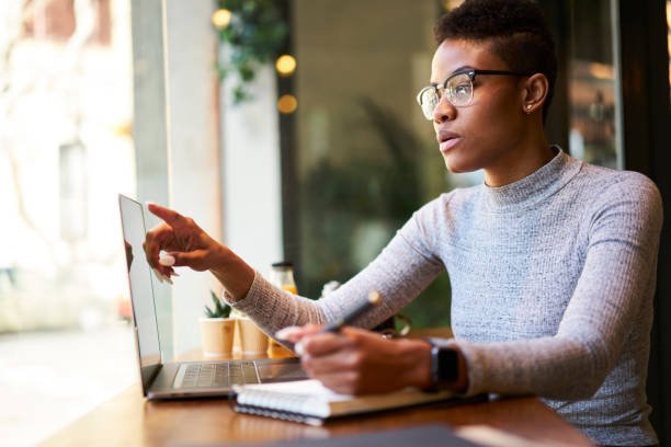 Concentrated attractive afro american female freelancer in casual outfit working with website designing writing down creative ideas into notebook to make concept planning sitting in coffee shop